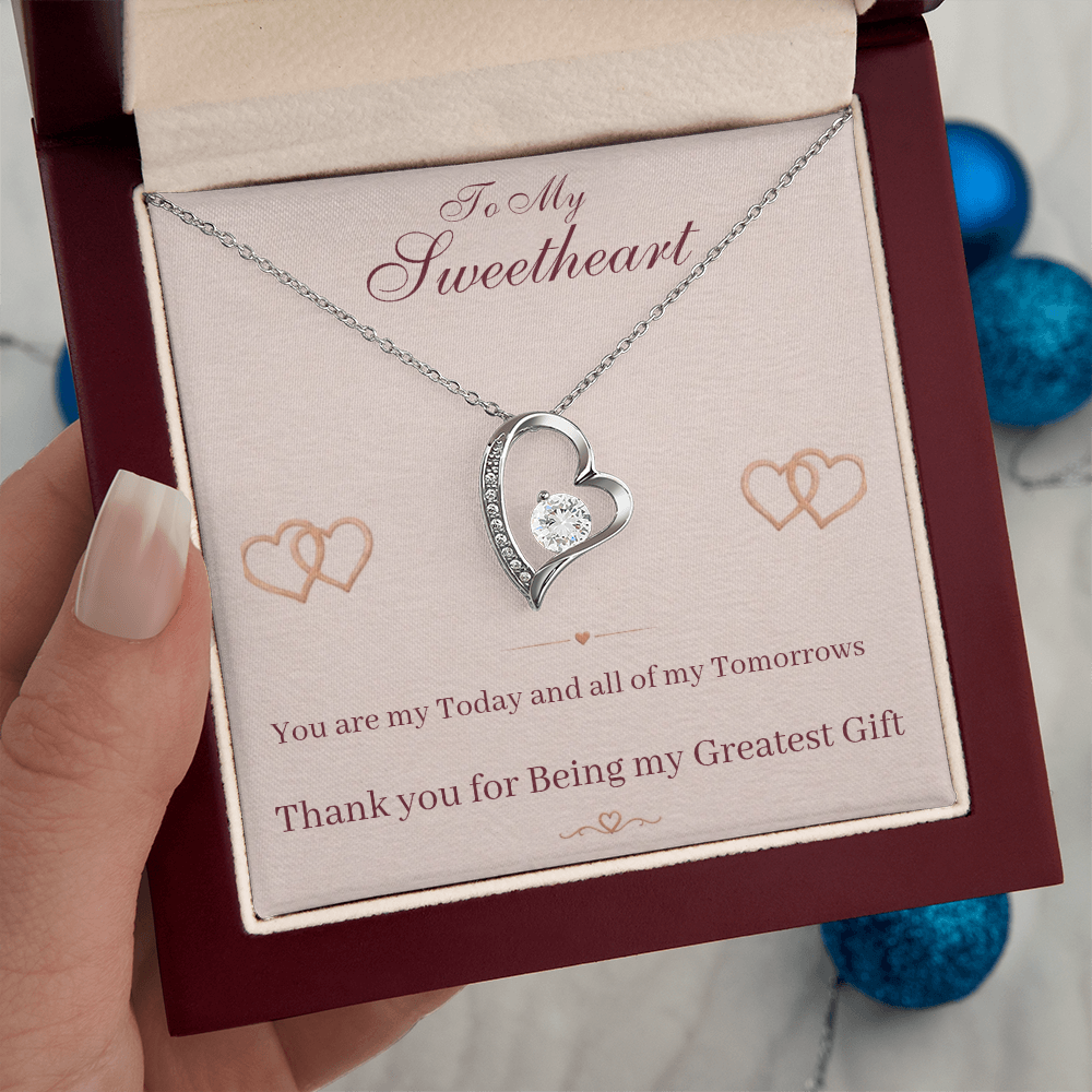Silver heart pendant necklace held above red gift box with sweetheart message card