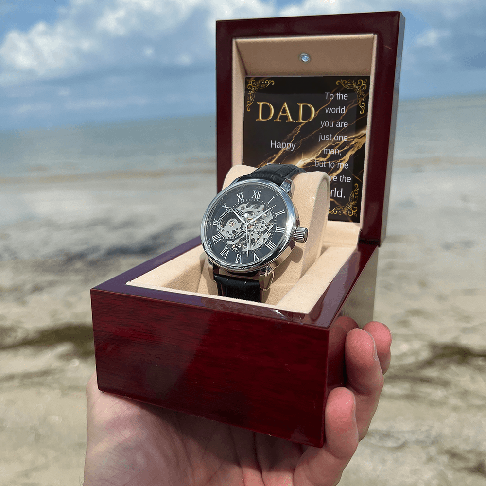 Wood watch box with DAD card and black leather open-work wristwatch on cream cushion
